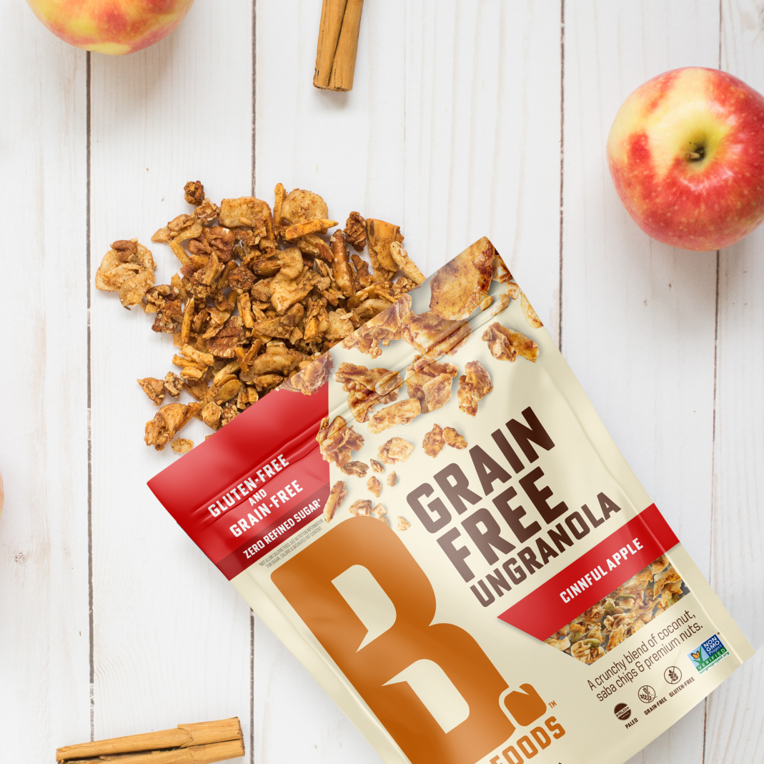 Bag of Cinnful Apple Grain Free Ungranola spilling out on white wood background with 2 apples and cinnamon sticks
