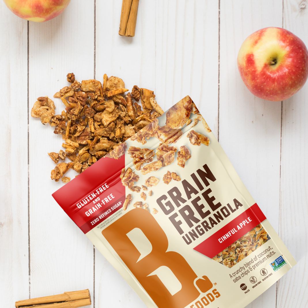 Bag of Cinnful Apple Grain Free Ungranola spilling out on white wood background with 2 apples and cinnamon sticks